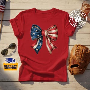 May include: A red short-sleeved t-shirt featuring a large central graphic of a bow patterned with the American flag, displaying blue fabric with white stars on the left side and red and white stripes on the right side of the bow design.
