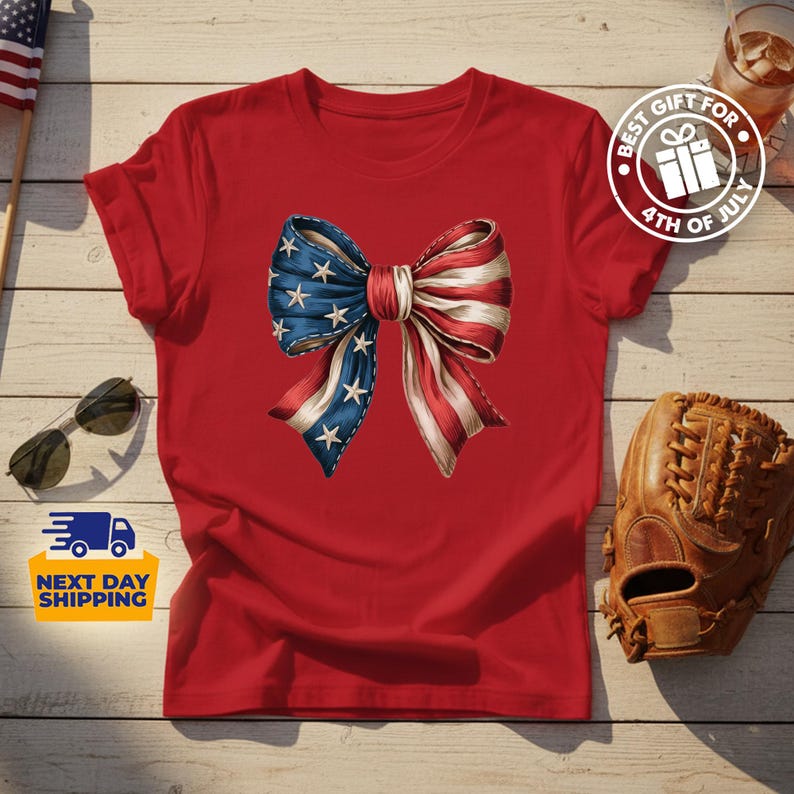 May include: A red short-sleeved t-shirt featuring a large central graphic of a bow patterned with the American flag, displaying blue fabric with white stars on the left side and red and white stripes on the right side of the bow design.
