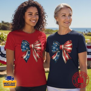 May include: A red short-sleeved t-shirt and a navy blue short-sleeved t-shirt, each featuring a large graphic of a bow decorated with a pattern of white stars on a blue background and red and white stripes, centered on the chest of each garment.