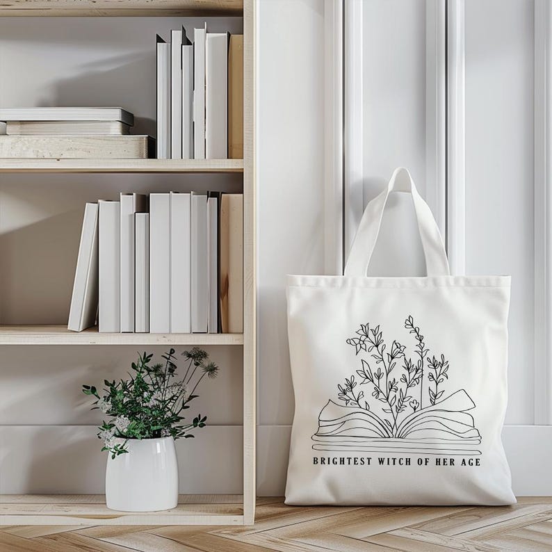 May include: White canvas tote bag with a black line drawing of an open book with flowers growing out of it. The text "Brightest Witch of Her Age" is printed below the book.