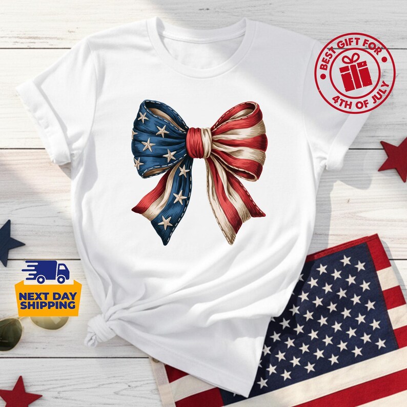 May include: A white short-sleeved t-shirt featuring a central graphic of a large bow with a pattern of blue with white stars on one side and red and white stripes on the other. The shirt includes a circular red badge with the text BEST GIFT FOR 4TH OF JULY.