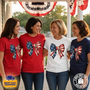 May include: A collection of short-sleeved t-shirts in red, white, and navy blue, each featuring a large, central graphic of a decorative bow patterned with the stars and stripes of the American flag. The shirts are worn by four individuals.