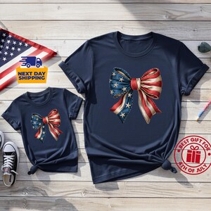 May include: Two navy blue short-sleeve t-shirts featuring a large graphic of a bow with a red, white, and blue American flag pattern. The bow design includes white stars on a blue background on one side and red and white stripes on the other.