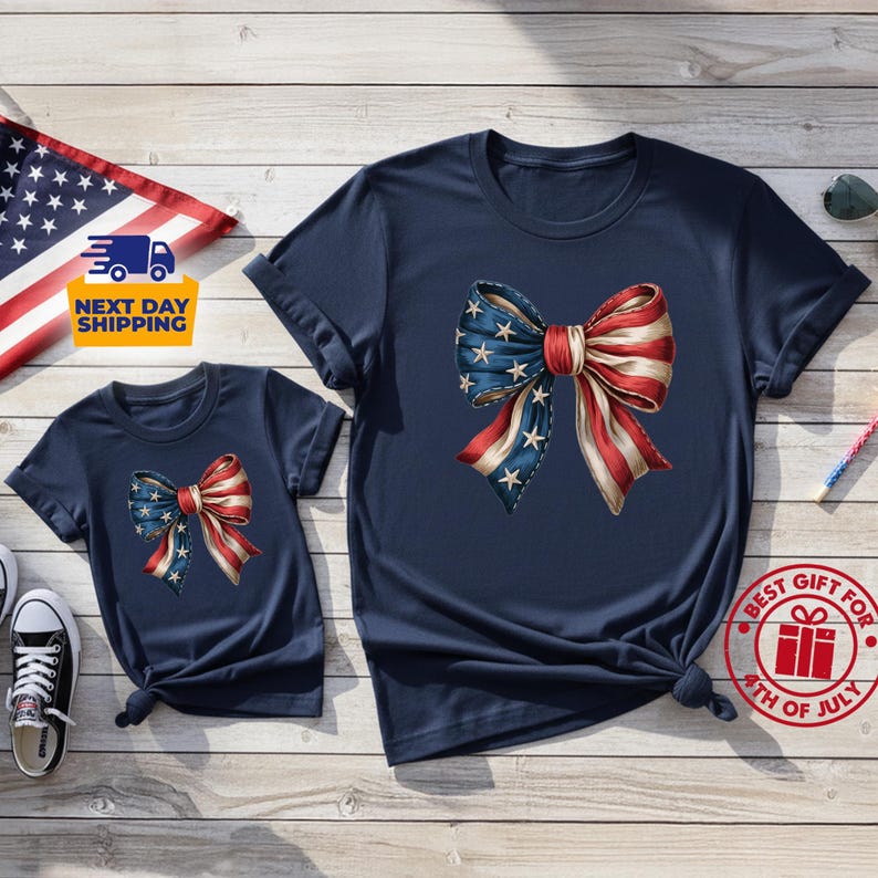 May include: Two navy blue short-sleeve t-shirts featuring a large graphic of a bow with a red, white, and blue American flag pattern. The bow design includes white stars on a blue background on one side and red and white stripes on the other.