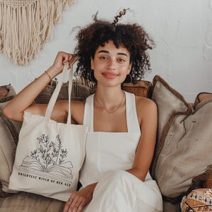May include: A white tote bag with black text and a floral design. The text reads "Brightest Witch of Her Age".