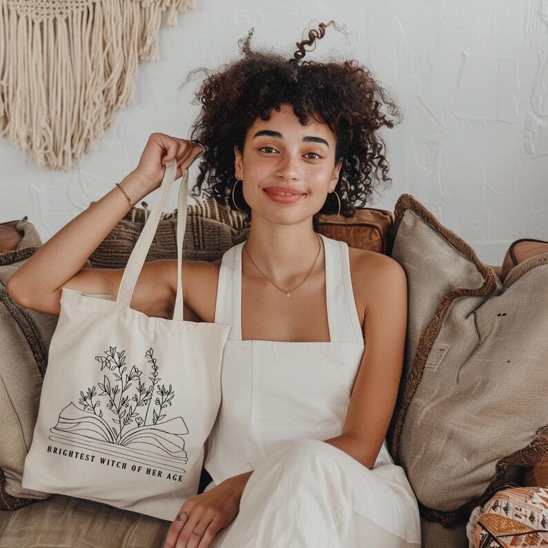May include: A white tote bag with black text and a floral design. The text reads "Brightest Witch of Her Age".