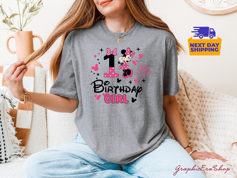K&ouml;nnte beinhalten: Graues T-Shirt mit einem Minnie-Mouse-Design, der Zahl 1, Herzen und dem Text "Birthday Girl" in Schwarz und Pink. Das Shirt hat einen Rundhalsausschnitt und kurze &Auml;rmel. Das Design beinhaltet eine Minnie-Mouse-Grafik.
