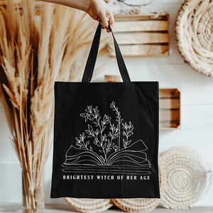 May include: Black canvas tote bag with a white outline of an open book with flowers growing from it. The text "Brightest Witch of Her Age" is printed below the book.