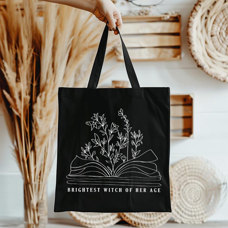 May include: Black canvas tote bag with a white outline of an open book with flowers growing from it. The text "Brightest Witch of Her Age" is printed below the book.