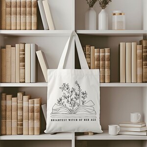 May include: White tote bag with a black line drawing of an open book with flowers growing out of it. The text "Brightest Witch of Her Age" is printed below the book.