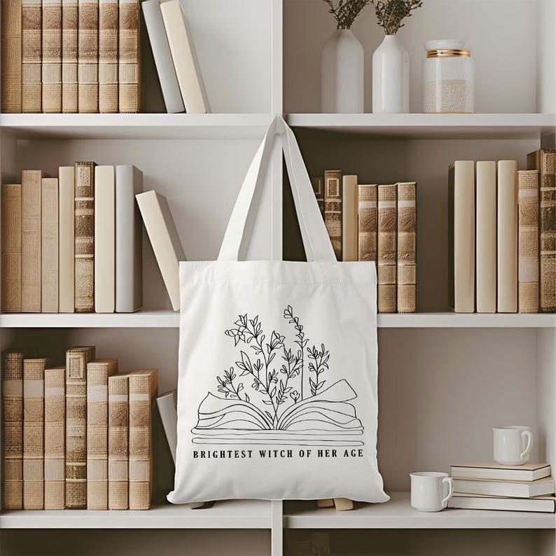 May include: White tote bag with a black line drawing of an open book with flowers growing out of it. The text "Brightest Witch of Her Age" is printed below the book.