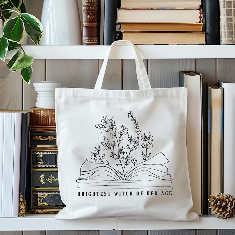 May include: White canvas tote bag with a black line drawing of an open book with flowers growing out of it. The text "Brightest Witch of Her Age" is printed below the book.