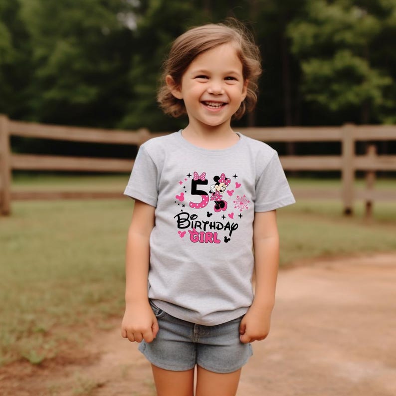 K&ouml;nnte beinhalten: Hellgraues T-Shirt mit dem Aufdruck "5 Birthday Girl". Das Design umfasst eine rosa Zahl 5, Minnie Mouse, rosa Herzen und Blumen. Das T-Shirt wird von einem jungen M&auml;dchen getragen.
