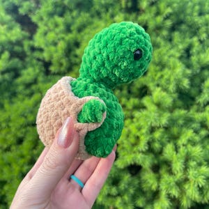May include: A handmade green and tan crochet turtle toy. The turtle has a green head and body, with a tan shell. The turtle has black eyes. The toy is held in a hand against a green leafy background.