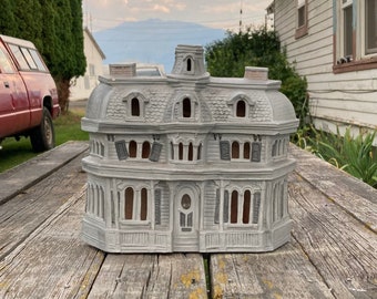 Puede incluir: Una casa en miniatura de cerámica gris claro con techo abuhardillado, múltiples ventanas arqueadas y contraventanas gris oscuro. La estructura tiene una puerta central, una pequeña torre en el techo y dos chimeneas con molduras detalladas.