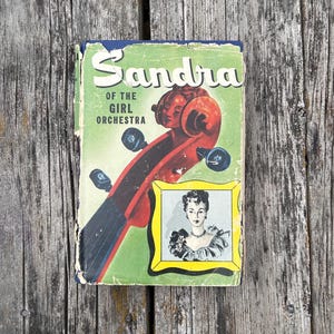 May include: A vintage hardback book titled Sandra of the Girl Orchestra with a green cover featuring a red violin scroll and a small framed illustration of a person. The book shows signs of wear along the edges and corners of the dust jacket.