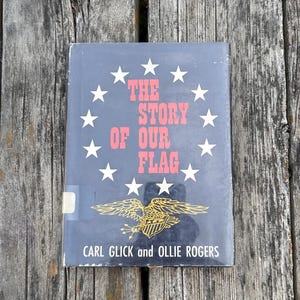 May include: A hardcover book titled The Story of Our Flag by Carl Glick and Ollie Rogers. The black cover features a circle of twelve white stars surrounding the red text, with a gold eagle illustration and a small white library sticker.