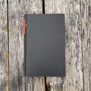 Pu&ograve; includere: Un libro rettangolare con copertina rigida nera e una sottile striscia verticale rossa lungo il bordo sinistro del dorso. Il libro &egrave; posizionato verticalmente e presenta una copertina pulita e semplice, priva di testo o grafiche visibili.