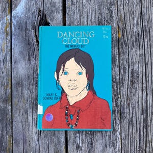 May include: A hardcover book titled Dancing Cloud The Navajo Boy by Mary and Conrad Buff. The cover features a teal background with a central illustration of a person with dark hair wearing a red shirt and a beaded necklace. Text includes 970.1 Bu Sw.