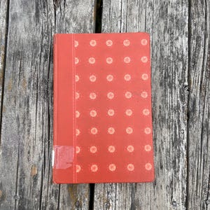 May include: A rectangular hardcover book with a vibrant orange cover featuring a repeating pattern of light-colored circular motifs. A vertical strip runs along the left side, and a small white rectangular label is attached to the bottom corner.