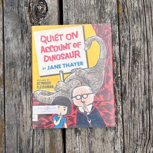 Pode incluir: Um livro infantil de capa dura intitulado Quiet on Account of Dinosaur de Jane Thayer com ilustra&ccedil;&otilde;es de Seymour Fleishman. A capa mostra um grande dinossauro castanho, uma rapariga e um homem de &oacute;culos a fazer um gesto de sil&ecirc;ncio.