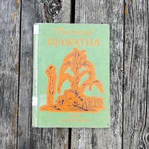 K&ouml;nnte beinhalten: Ein gebundenes Buch mit dem Titel The Story of HIAWATHA, adaptiert von Allen Chappel. Der Einband ist hellgr&uuml;n mit orangefarbenen Illustrationen einer Person neben einem gro&szlig;en Maiskolben und einer Menschengruppe. Der Titel ist orange.