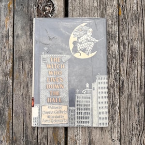 Puede incluir: Un libro de tapa dura titulado The Witch Who Lives Down the Hall, escrito por Donna Guthrie e ilustrado por Amy Schwartz. La portada presenta una ilustraci&oacute;n en blanco y negro de un paisaje urbano y una bruja en una escoba ante una luna amarilla.