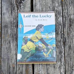 Pu&ograve; includere: Un libro con copertina rigida intitolato Leif the Lucky di Erick Berry, illustrato da William Plummer. La copertina presenta uno sfondo blu con una figura vestita di giallo, una nave vichinga e una grafica a rosa dei venti. &Egrave; un'edizione Discovery Book.
