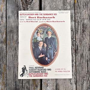 K&ouml;nnte beinhalten: Ein Notenheft f&uuml;r den Film Butch Cassidy und Sundance Kid, komponiert von Burt Bacharach. Das Cover zeigt ein zentrales ovales Portr&auml;t von Paul Newman, Robert Redford und Katharine Ross, erg&auml;nzt durch roten und schwarzen Text.