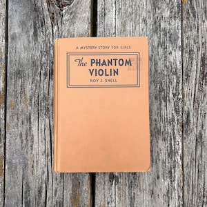 K&ouml;nnte beinhalten: Ein gebundenes Buch mit dem Titel The Phantom Violin von Roy J. Snell und dem Untertitel A Mystery Story for Girls. Das Buch hat einen einfarbigen, hellbraunen Einband mit schwarzem Text in einem rechteckigen Rahmen auf der Vorderseite.