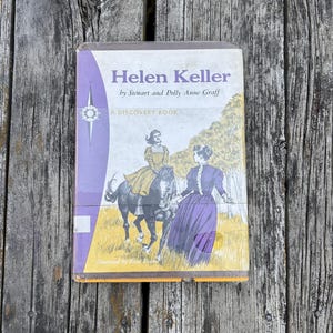 May include: A hardback book titled Helen Keller by Stewart and Polly Anne Graff, A Discovery Book. The cover features a vintage illustration of a person riding a black horse and another person standing in a field, with purple and yellow accents.