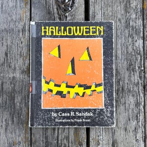 May include: A hardback book titled HALLOWEEN by Cass R. Sandak with illustrations by Frank Bozzo. The cover features a central orange jack-o'-lantern graphic with yellow triangular eyes and a jagged yellow mouth set against a black background.