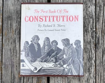 Może przedstawiać: Książka w twardej oprawie zatytułowana The First Book of The Constitution autorstwa Richarda B. Morrisa z ilustracjami Leonarda Everetta Fishera. Biała okładka zawiera czarno-białą ilustrację postaci historycznych podpisujących dokument.
