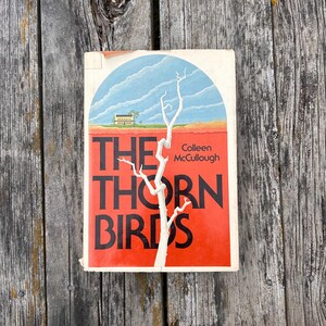 May include: A paperback book titled The Thorn Birds by Colleen McCullough. The cover features a white, leafless tree against a blue sky and a red landscape with a small house. The title is printed in large black letters over the red background.