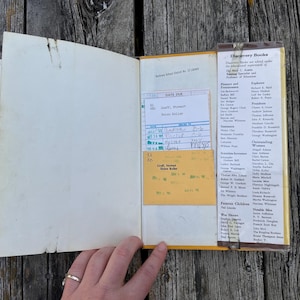 May include: An open hardback book with a white endpaper and a yellow library checkout card pocket. The pocket contains a white date due slip with handwritten entries. A list of Discovery Books is printed on the right side of the inner cover.