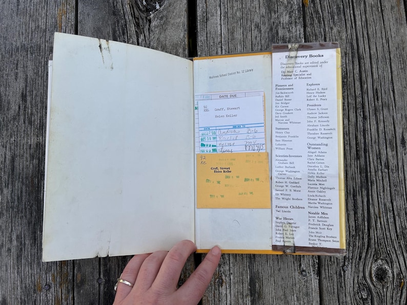May include: An open hardback book with a white endpaper and a yellow library checkout card pocket. The pocket contains a white date due slip with handwritten entries. A list of Discovery Books is printed on the right side of the inner cover.