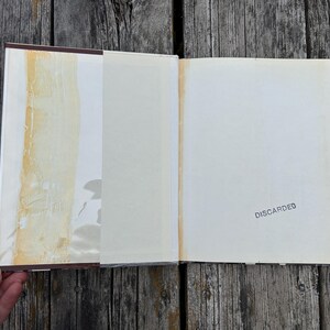 Puede incluir: Un libro de tapa dura abierto con guardas blancas. La p&aacute;gina izquierda presenta una franja vertical de residuos adhesivos amarillentos. La p&aacute;gina derecha contiene la palabra DISCARDED estampada con tinta negra.