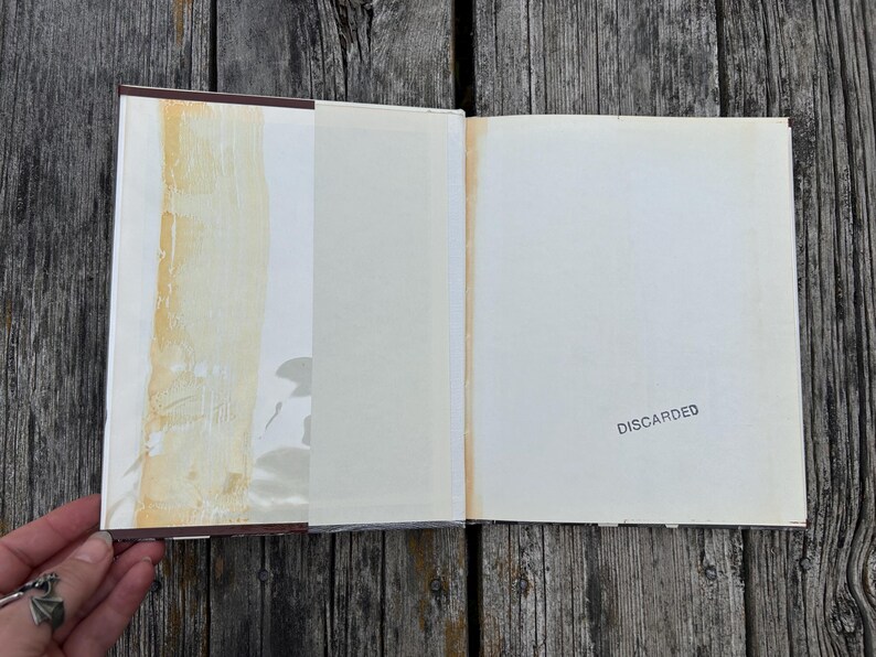 Puede incluir: Un libro de tapa dura abierto con guardas blancas. La p&aacute;gina izquierda presenta una franja vertical de residuos adhesivos amarillentos. La p&aacute;gina derecha contiene la palabra DISCARDED estampada con tinta negra.