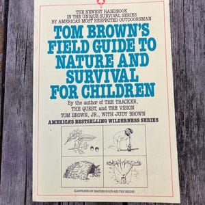 Puede incluir: Un libro antiguo titulado "Tom Brown's Field Guide to Nature and Survival for Children". La portada presenta un fondo crema con texto azul y marrón, e ilustraciones de técnicas de supervivencia. El libro forma parte de la "America's Bestselling Wilderness Series".