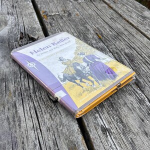 May include: A paperback book titled Helen Keller by Stewart and Polly Anne Graff, featuring a cover illustration of a person riding a black horse and another person standing nearby, encased in a clear plastic protective cover with purple accents.