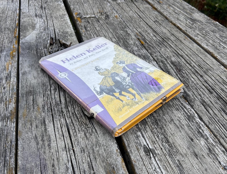 May include: A paperback book titled Helen Keller by Stewart and Polly Anne Graff, featuring a cover illustration of a person riding a black horse and another person standing nearby, encased in a clear plastic protective cover with purple accents.