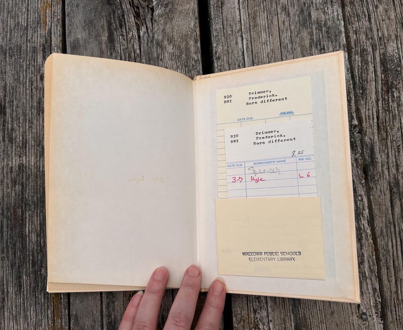 Pu&ograve; includere: Un libro con copertina rigida aperto con una pagina color crema e una tasca per la scheda della biblioteca. La scheda mostra il testo Brimmer, Frederick, Born different, Wallowa Public Schools Elementary Library e nomi scritti a mano.