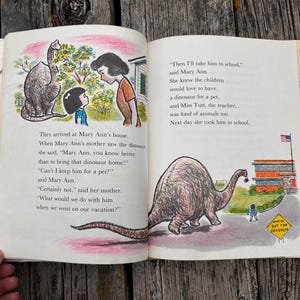 Pode incluir: Um livro infantil vintage aberto com duas p&aacute;ginas de texto e ilustra&ccedil;&otilde;es coloridas. A p&aacute;gina esquerda mostra uma menina e uma mulher falando com um dinossauro. A p&aacute;gina direita mostra um grande dinossauro caminhando em dire&ccedil;&atilde;o a uma escola.