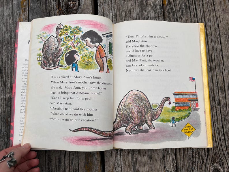 Pode incluir: Um livro infantil vintage aberto com duas p&aacute;ginas de texto e ilustra&ccedil;&otilde;es coloridas. A p&aacute;gina esquerda mostra uma menina e uma mulher falando com um dinossauro. A p&aacute;gina direita mostra um grande dinossauro caminhando em dire&ccedil;&atilde;o a uma escola.