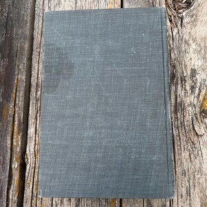 May include: A rectangular hardcover book with a solid dark gray, textured fabric cover. The book is positioned vertically, displaying a plain front cover with a visible weave pattern in the material and slightly worn edges along the spine.