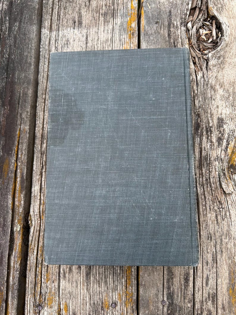 May include: A rectangular hardcover book with a solid dark gray, textured fabric cover. The book is positioned vertically, displaying a plain front cover with a visible weave pattern in the material and slightly worn edges along the spine.