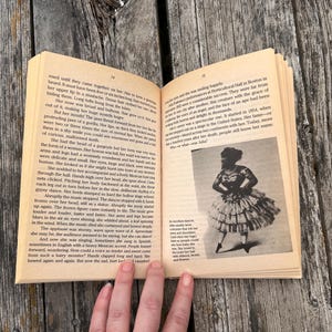 Pu&ograve; includere: Un libro aperto con pagine color beige contenente testo stampato e una fotografia in bianco e nero di una persona con un abito a balze. La pagina sinistra presenta diversi paragrafi, mentre quella destra include testo e l'immagine di un artista.
