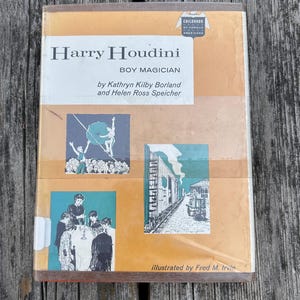 May include: A hardcover book titled Harry Houdini Boy Magician by Kathryn Kilby Borland and Helen Ross Speicher. The tan and white cover features three illustrations depicting a tightrope walker, a group of children, and a train car scene.