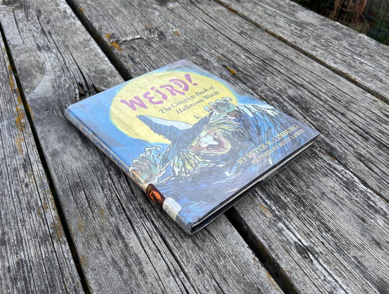 以下が含まれることがあります： ピーター・R・リンバーグ著、ベッツィー・リューイン絵のハードカバー本『Weird! The Complete Book of Halloween Words』。表紙には大きな黄色い月と、とんがり帽子をかぶった魔女のカラフルなイラストが描かれています。