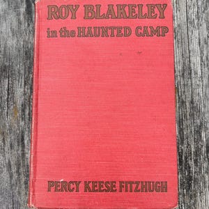 May include: A red hardcover book titled Roy Blakeley in the Haunted Camp by Percy Keese Fitzhugh. The front cover features dark brown text printed on a textured red fabric surface, with the title at the top and the author name at the bottom.
